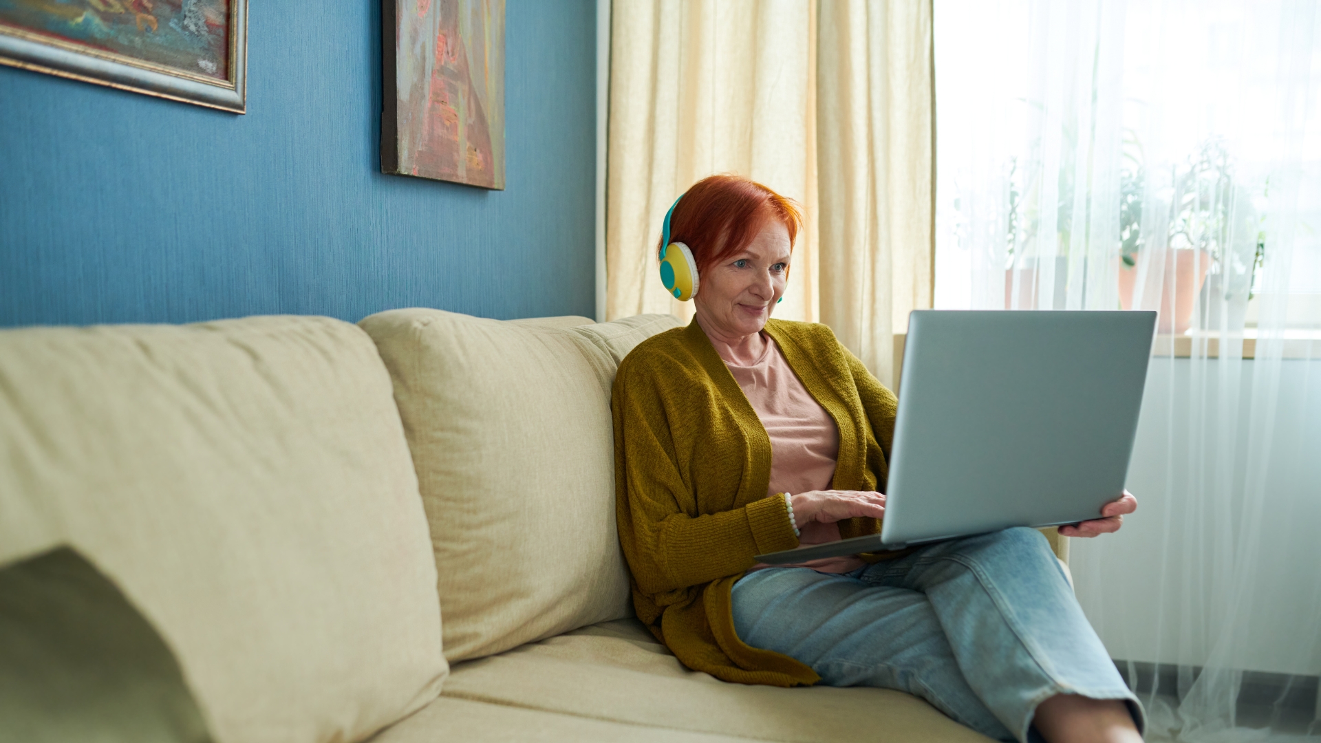 Kuvassa vanhempi nainen istuu sohvalla kuulokkeet paassa ja kannettava tietokone sylissa, liittyy Kesan tunnelmaa etalukupiiriin eopiston tarjonnassa.